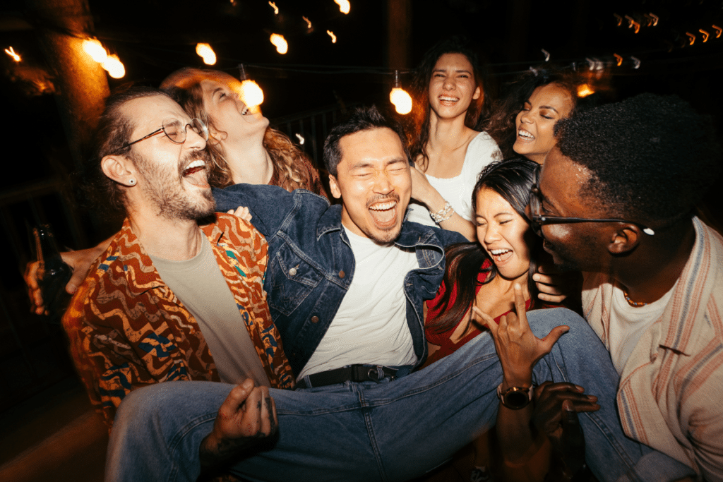 um homem sorrindo enquanto é carregado por um grupo de amigos em uma festa
